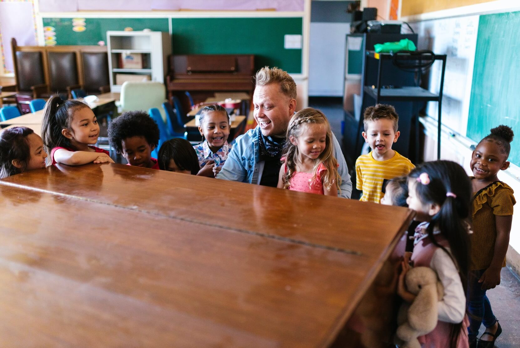 Lots of children and one male teacher gathered around a piano singing